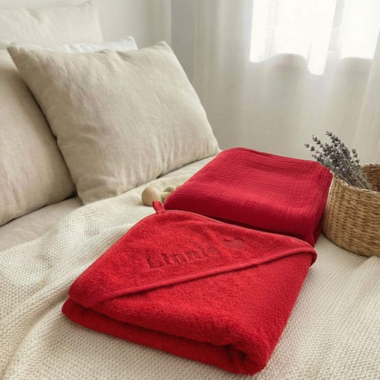 Hier liegt das Geburts-Set bestehend aus einem roten Geburtstuch und einem  roten Baby Kapuzenhandtuch nebeneinander auf dem Bett präsentiert