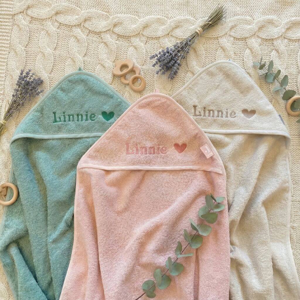 Baby Kapuzenhandtücher in rosa, grün und beige liegen ausgebreitet und mit Namen und Herz personalisiert auf einer weichen Decke