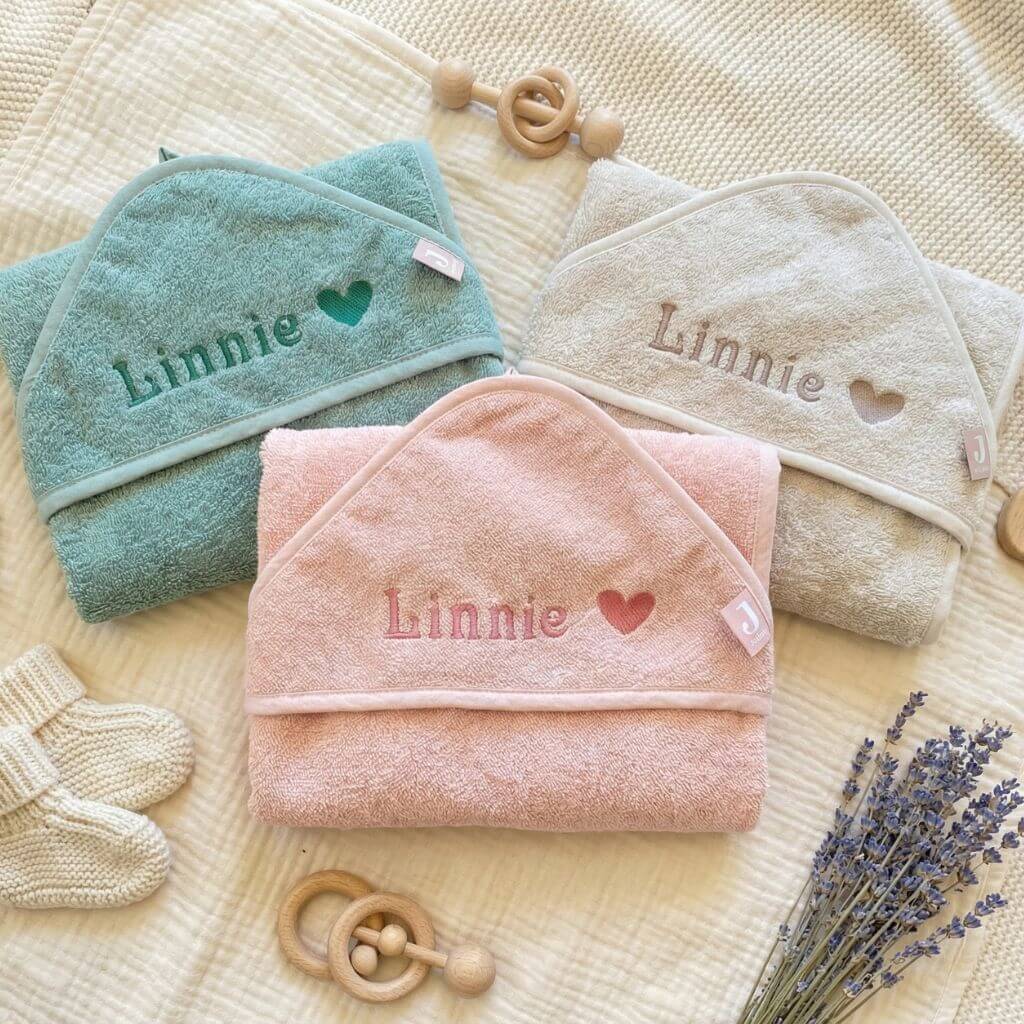Baby Kapuzenhandtücher in rosa, grün und beige liegen zusammengefaltet mit Namen und Herz personalisiert auf einer Decke, darum liegen Holz-Baby Spielsachen und Lavendel
