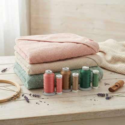 Baby Kapuzenhandtücher in rosa, beige und grün auf einem Stapel und davor sind die farblich passenden Stickgarnrollen für die Personalisierung aufgestellt