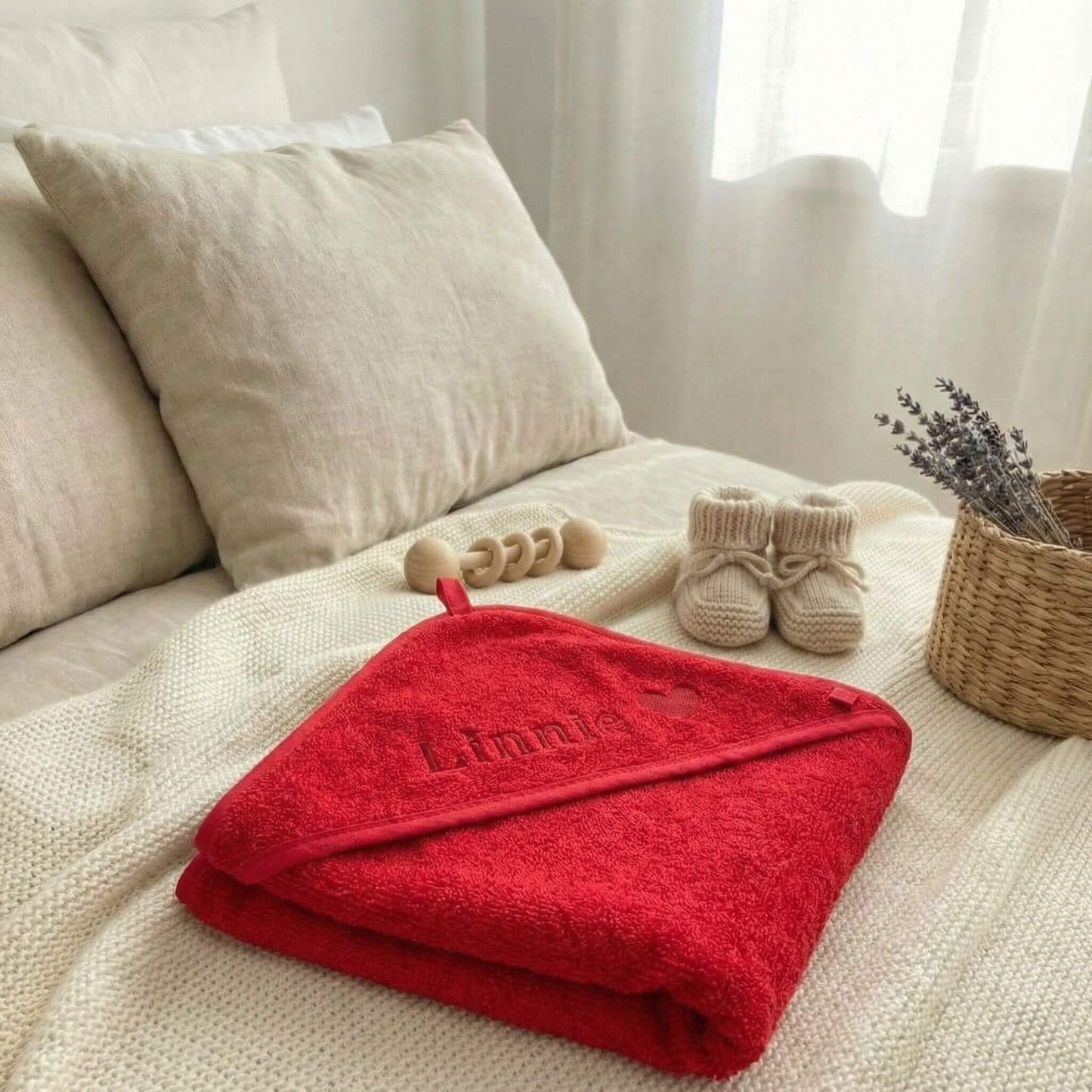 Rotes Baby Kapuzenhandtuch liegt zusammengefaltet mit Namen und Herz bestickt auf dem Bett